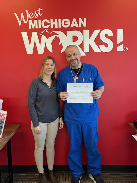 Man in scrubs holding a certificate standing next to a woman against a red background with the West Michigan Works! logo behind them