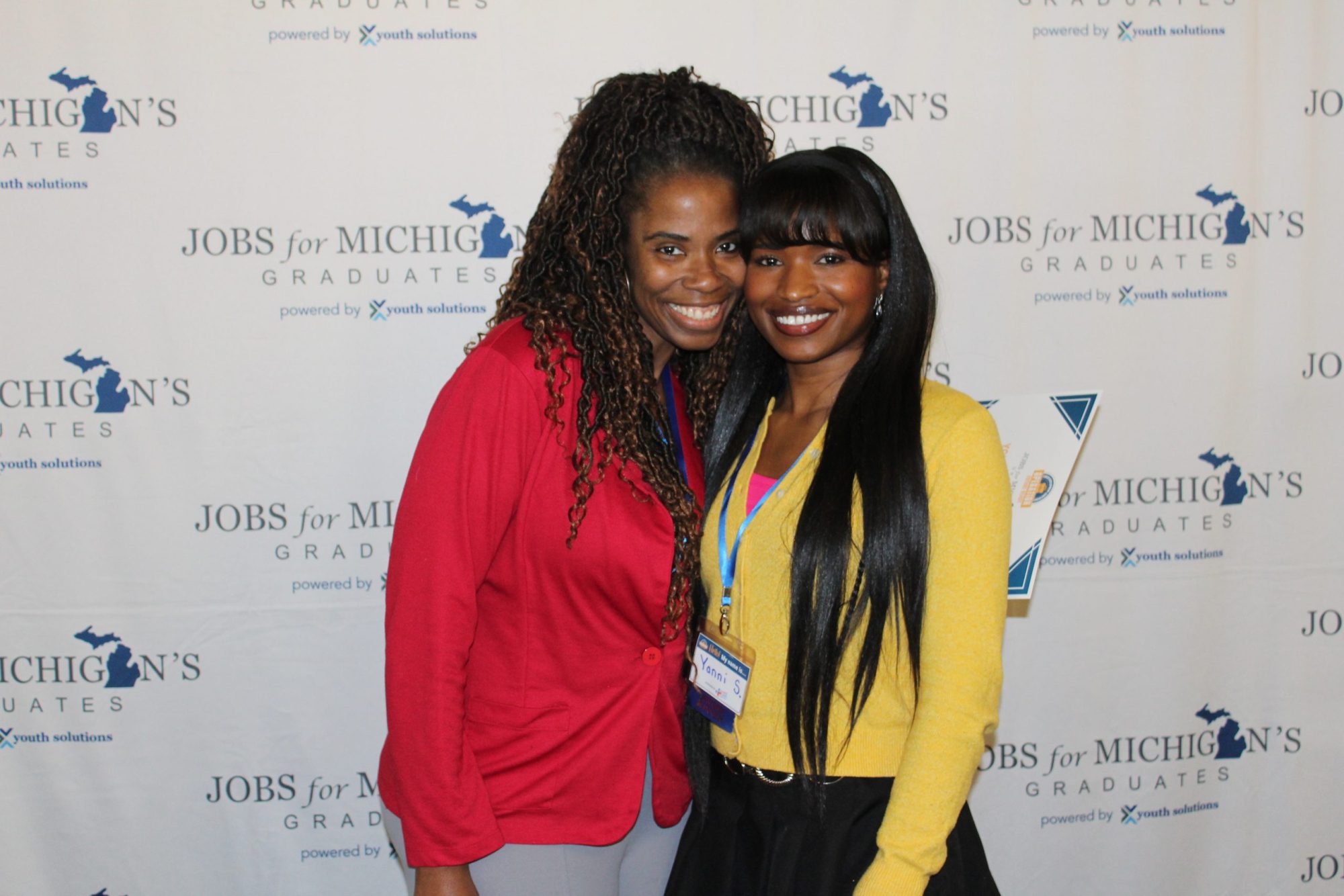 Two women posing together in front of a Jobs for Michigan Graduates backdrop